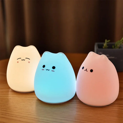 Three silicone cat night lights glowing cream, blue and pink on a wooden table