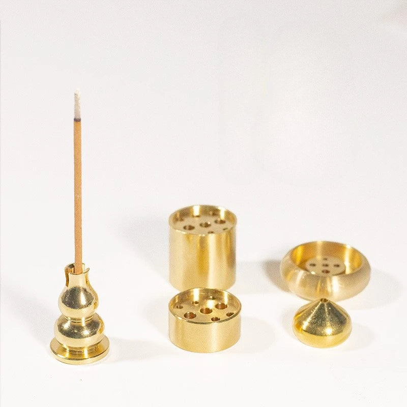 Three of the gold-coloured metal incense holders together - a gourd post, a drilled barrel and a low cone - with a single incense stick standing in one. The incense is not included