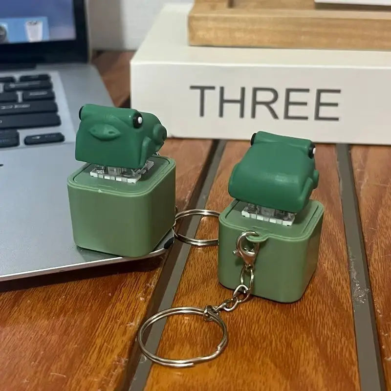 Two green frog keycap keychains standing on a wooden desk beside a laptop keyboard