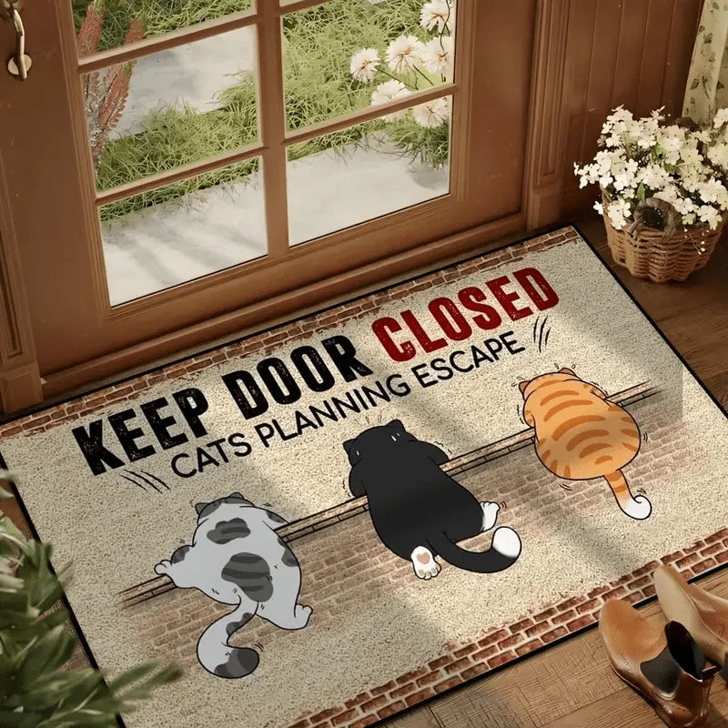 Doormat printed with three cats sneaking along a brick wall under the words KEEP DOOR CLOSED CATS PLANNING ESCAPE, laid inside a glazed door
