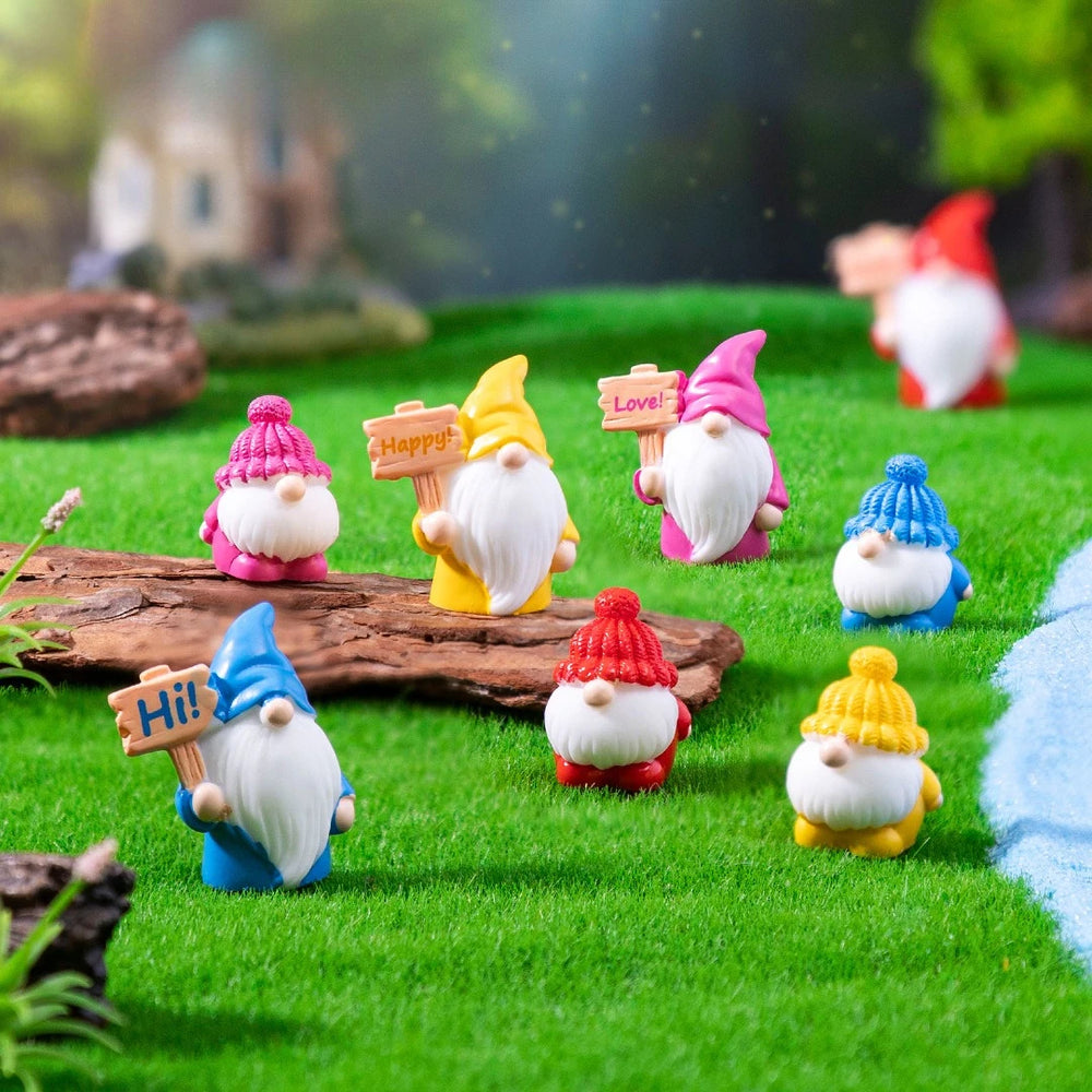 Eight tiny resin gnomes with pointed and knitted hats scattered on a moss mat, several holding little wooden signs reading Hi, Love and Happy - the moss, log and pond are staging and are not included