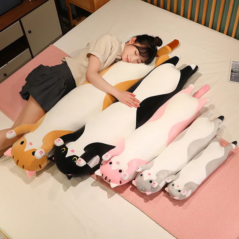 Five long cat body pillows in different colours laid out on a bed beside a person napping