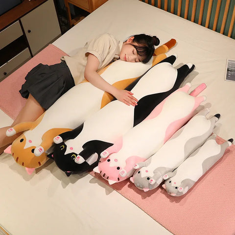 Five long cat body pillows in different colours laid out on a bed beside a person napping