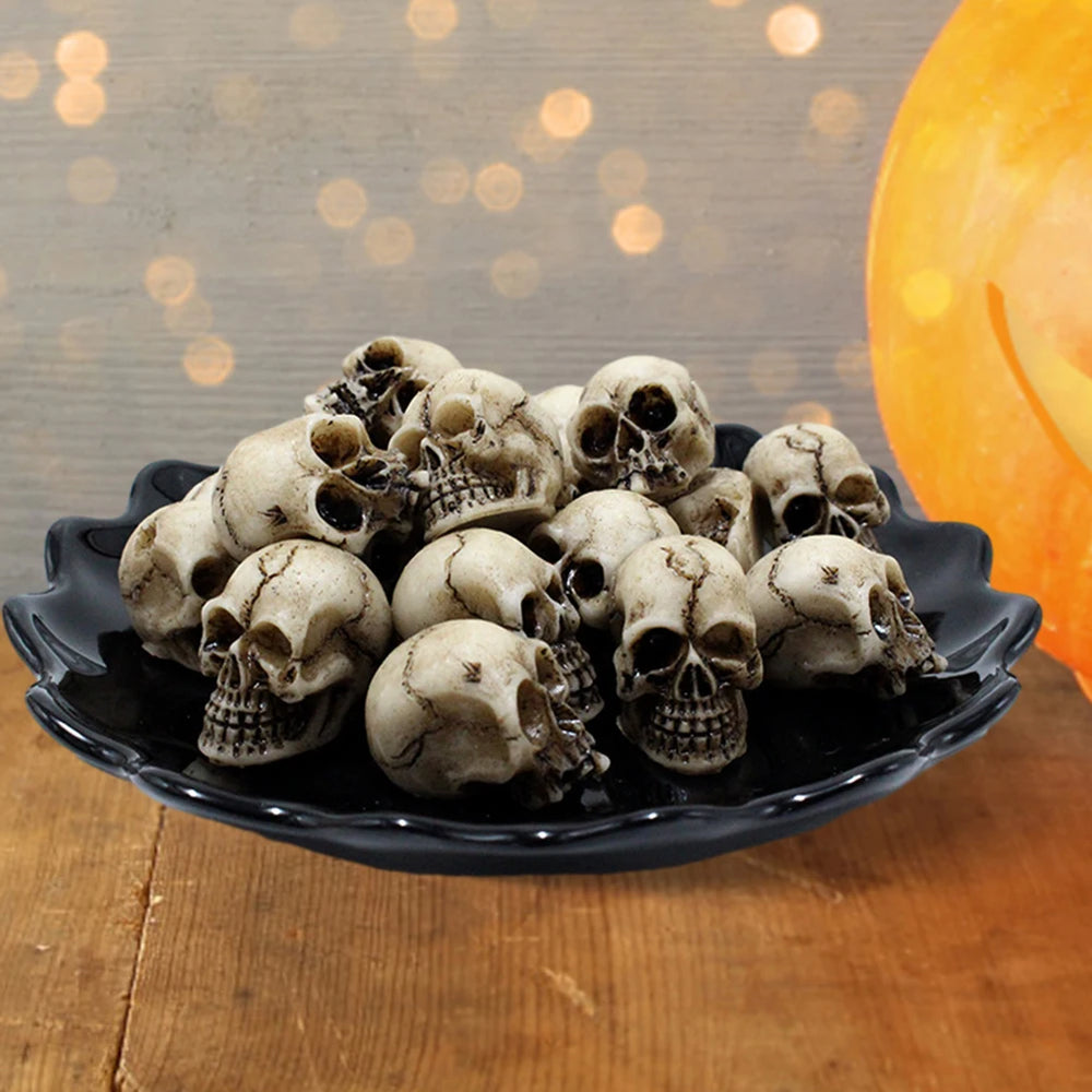 Twenty of the resin skulls piled in a black dish with a pumpkin lamp glowing behind - the dish and lamp are staging