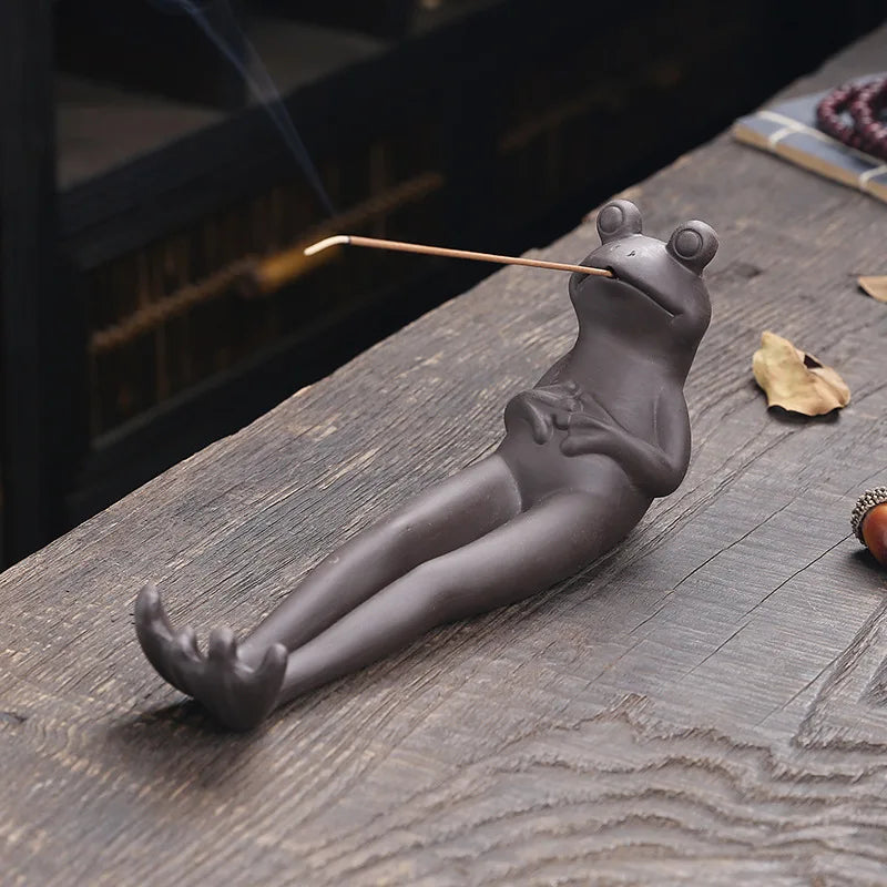Dark ceramic frog lying on its back holding a lit incense stick in its mouth