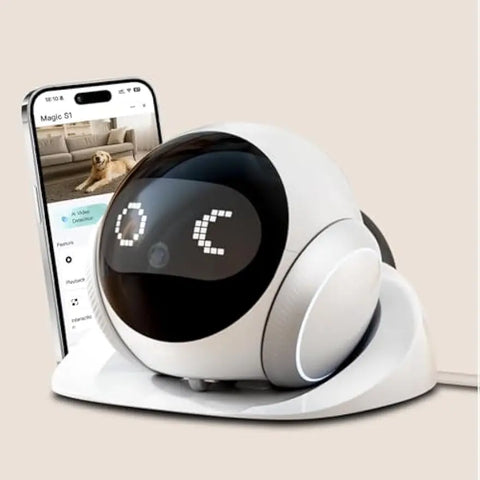 White ball-shaped robot with a black domed face showing dot-matrix eyes, on its charging dock beside a phone displaying a video feed