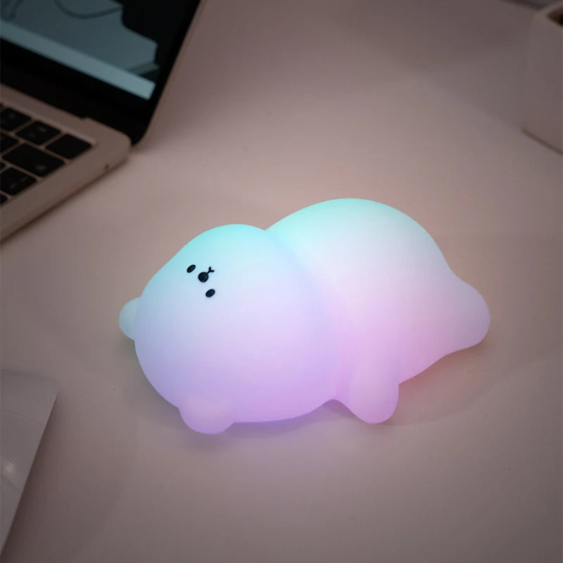 The plain lying-flat bear version on a desk beside a laptop, glowing in shifting green, pink and blue