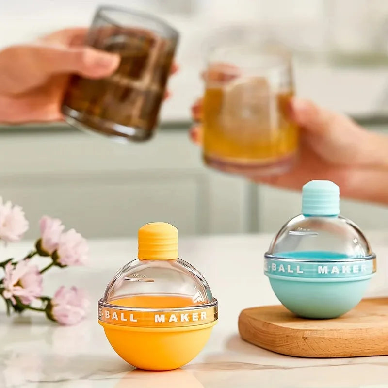 A yellow and a blue ice ball mold on a counter while two hands hold whisky glasses behind them