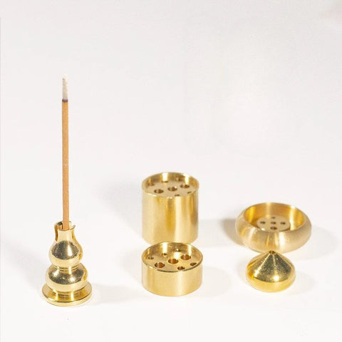 Three of the gold-coloured metal incense holders together - a gourd post, a drilled barrel and a low cone - with a single incense stick standing in one. The incense is not included