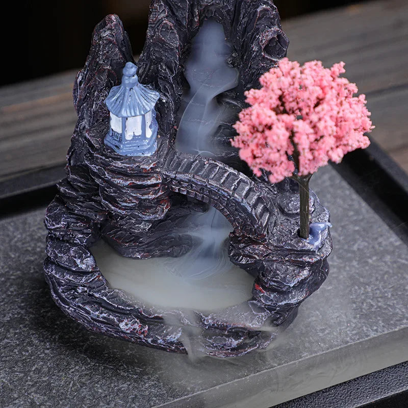 Resin mountain backflow incense burner with pink blossom tree and smoke pooling in the basin