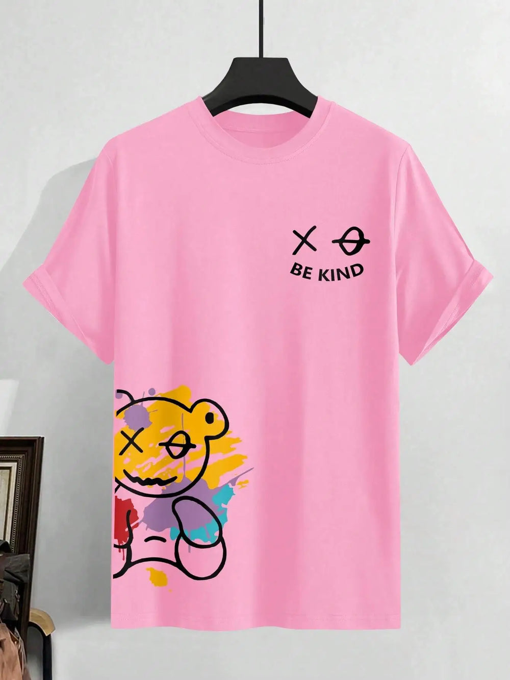 Pink cotton tee laid flat showing the BE KIND chest mark and the paint-splashed bear