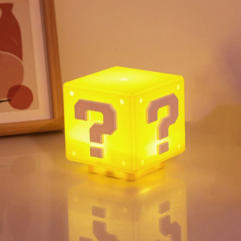 The yellow question mark cube lamp lit and glowing warm on a pale surface at night, reflected below, with a picture frame behind it