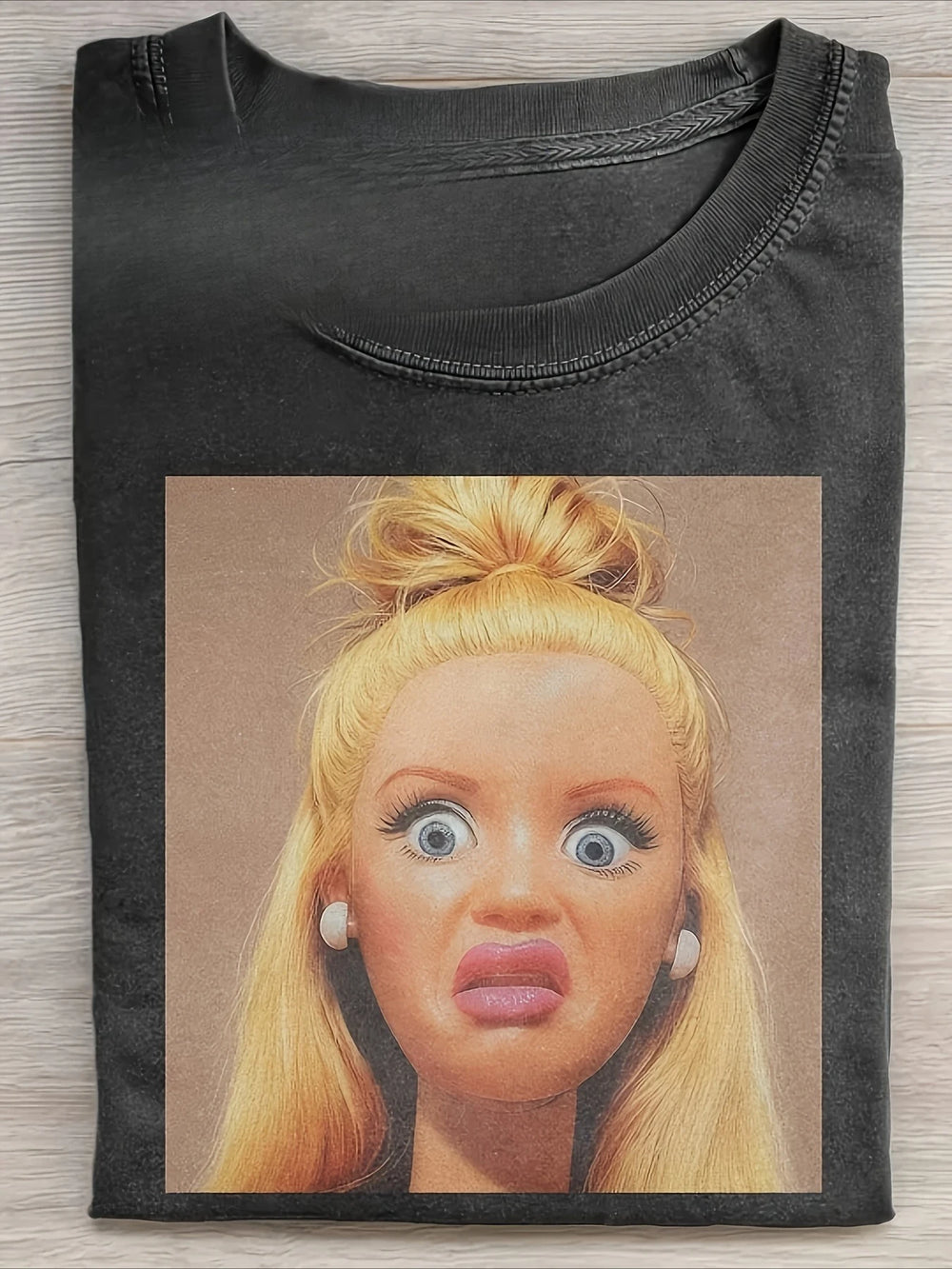 Washed black tee folded flat, printed with a photograph of a blonde fashion doll pulling a startled face