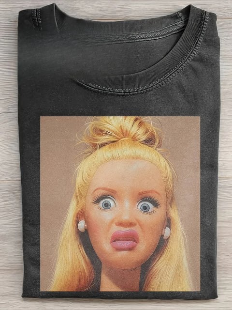 Washed black tee folded flat, printed with a photograph of a blonde fashion doll pulling a startled face