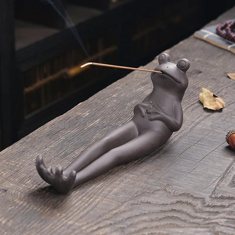Dark ceramic frog lying on its back holding a lit incense stick in its mouth