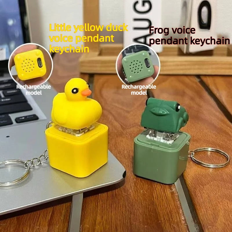 A yellow duck keycap keychain and a green frog keycap keychain side by side on a desk beside a laptop