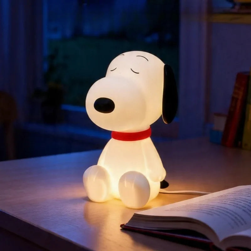A sitting cartoon dog lamp glowing warm white on a desk beside an open book at night, its cable running off to the right