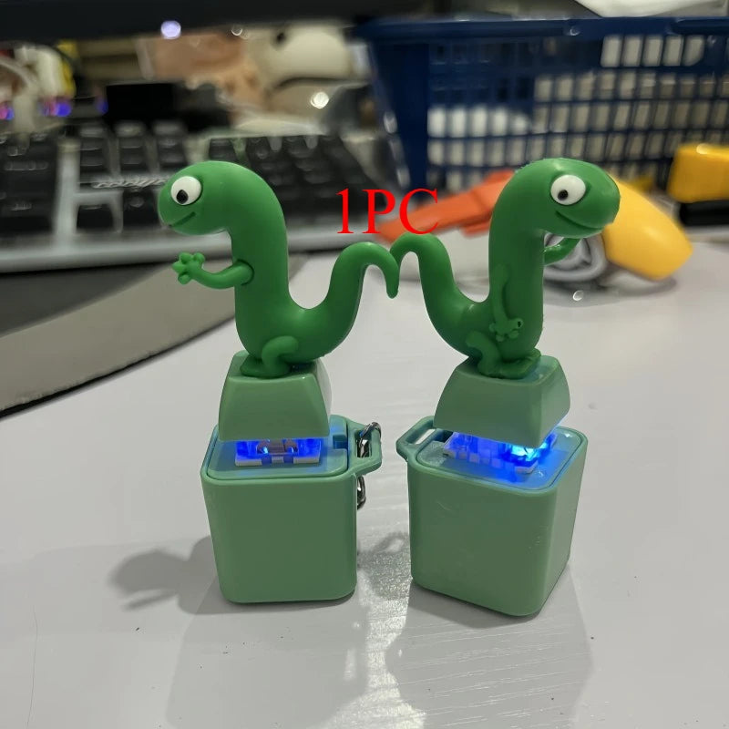 Two green lizard keycap keychains with their blue LEDs lit, standing on a desk